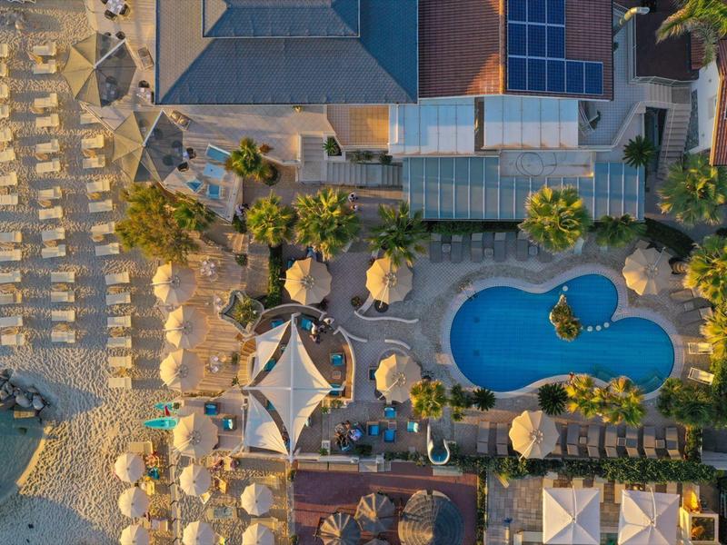 Aerial view of a beach resort with umbrellas, palm trees, a pool, and buildings.