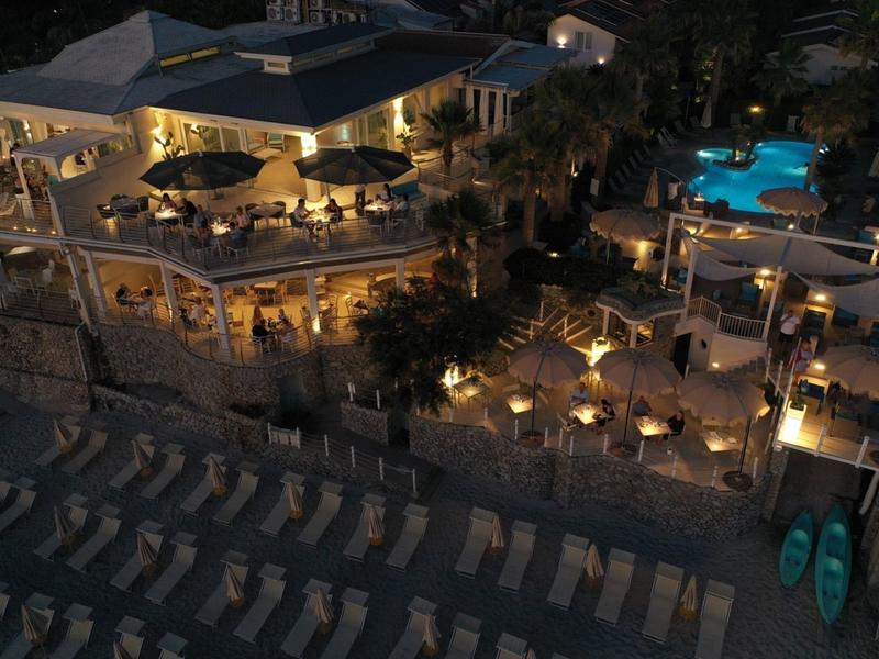 Night view of a lit resort with pool, outdoor dining, and empty beach chairs by the shore.