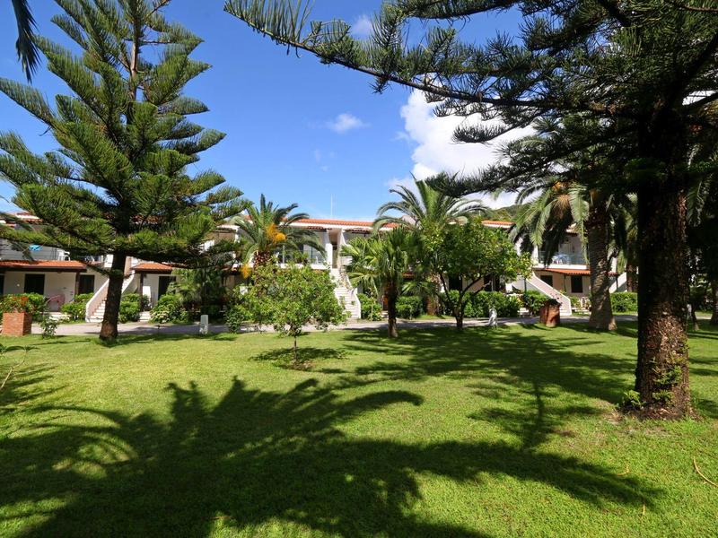 Jardin vert avec des palmiers et d'autres arbres devant un bâtiment hôtelier sous un ciel bleu.