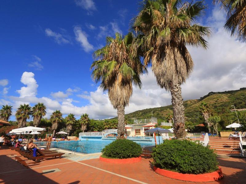 Espace piscine d'hôtel avec palmiers, chaises longues et montagnes en arrière-plan par temps ensoleillé.