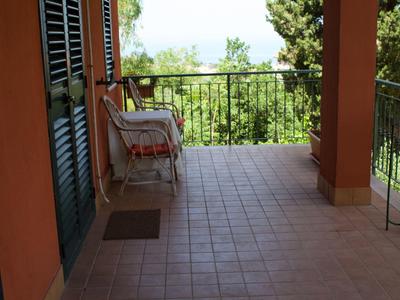 Große Terrasse mit braunen Fliesen, rotem Stuhl und Blick auf grünen Garten bei Sonnenschein.