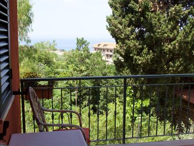 Balkon mit Metallgeländer, Tisch, Stuhl und Blick auf grüne Bäume und Gebäude im Hintergrund.
