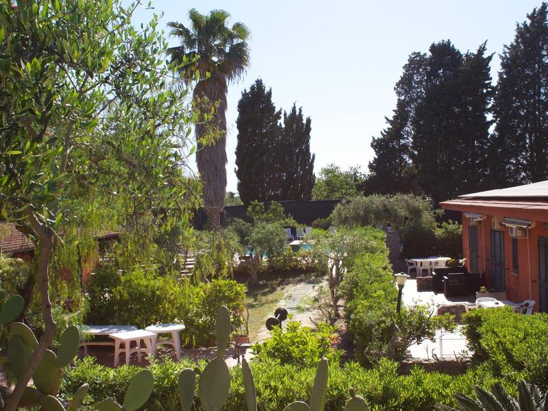 Grüner Garten mit Tisch, Bänken, Palmen und Zypressen neben rotem Haus unter klarem Himmel.