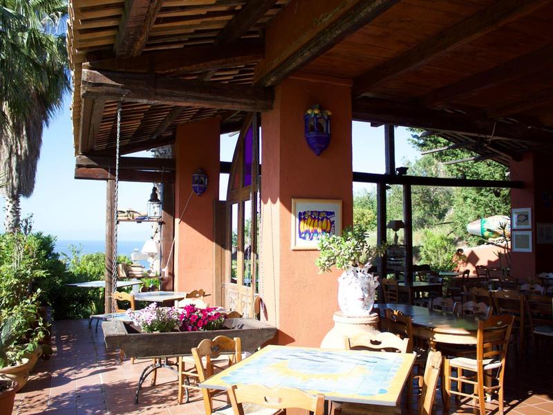 Gemütliche Terrasse mit Holzstühlen und Tischen, blühenden Pflanzen und Blick aufs Meer.