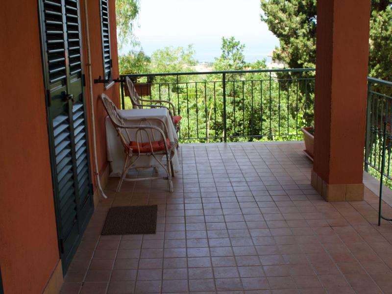Große Terrasse mit braunen Fliesen, rotem Stuhl und Blick auf grünen Garten bei Sonnenschein.