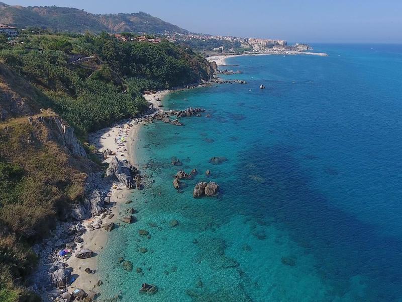 Côte avec eau bleue claire, rochers et collines verdoyantes en arrière-plan.
