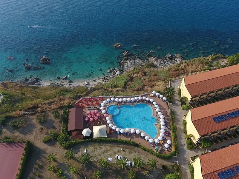 Vue aérienne d'un hôtel avec piscine à côté d'une côte claire et de la mer bleue.