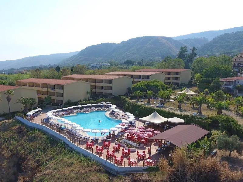 Complexe hôtelier avec piscine extérieure et terrasse ensoleillée sur une colline avec vue sur les montagnes.