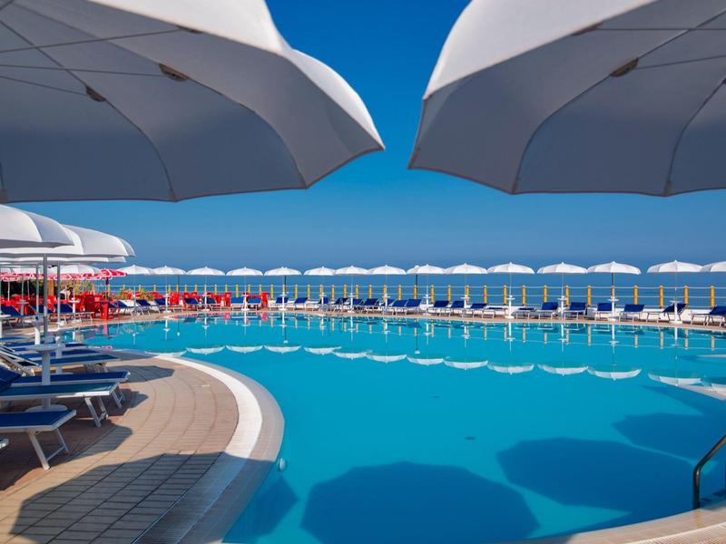 Grande piscine extérieure avec eau bleue et nombreux parasols blancs autour.