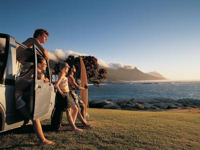 Drei Personen mit Surfboards stehen auf einer Wiese neben dem Meer bei Sonnenuntergang.