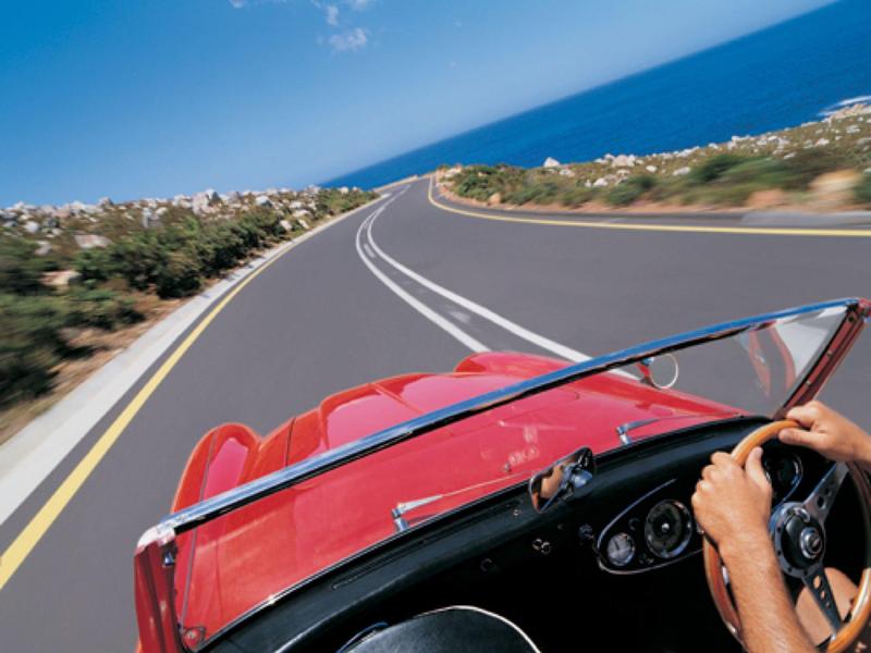Fahrer in rotem Cabrio auf kurviger Straße mit Meerblick bei sonnigem Wetter.