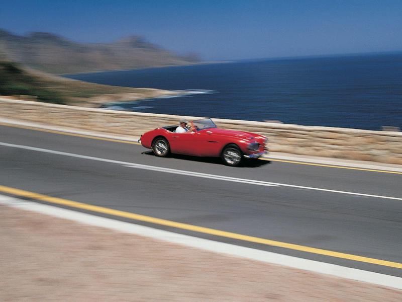Rotes Oldtimer-Cabrio fährt schnell auf Küstenstraße mit Meer und Bergen im Hintergrund.