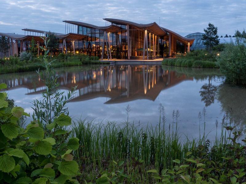 Modern hotelgebouw met glazen gevel en houten dak weerspiegelt in een vijver bij schemering.