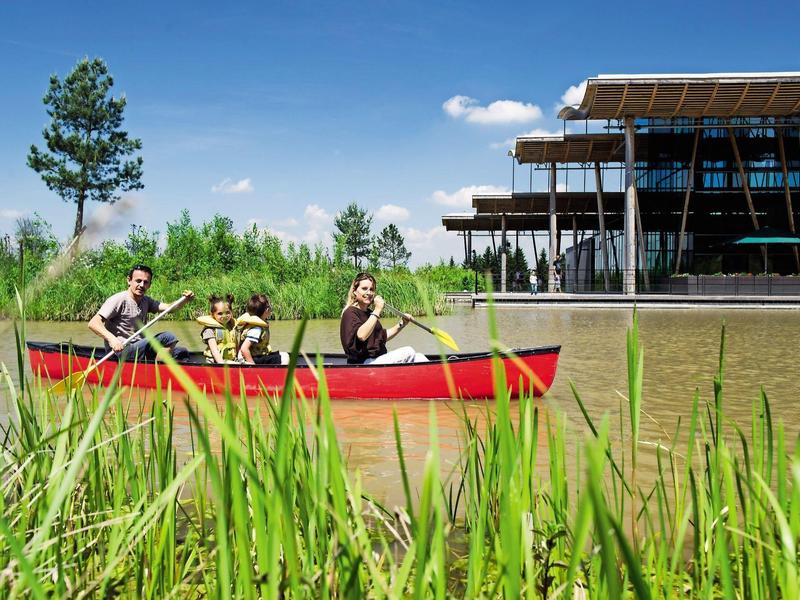 Familie peddelt in rode kano op rustig water nabij modern gebouw en bomen.