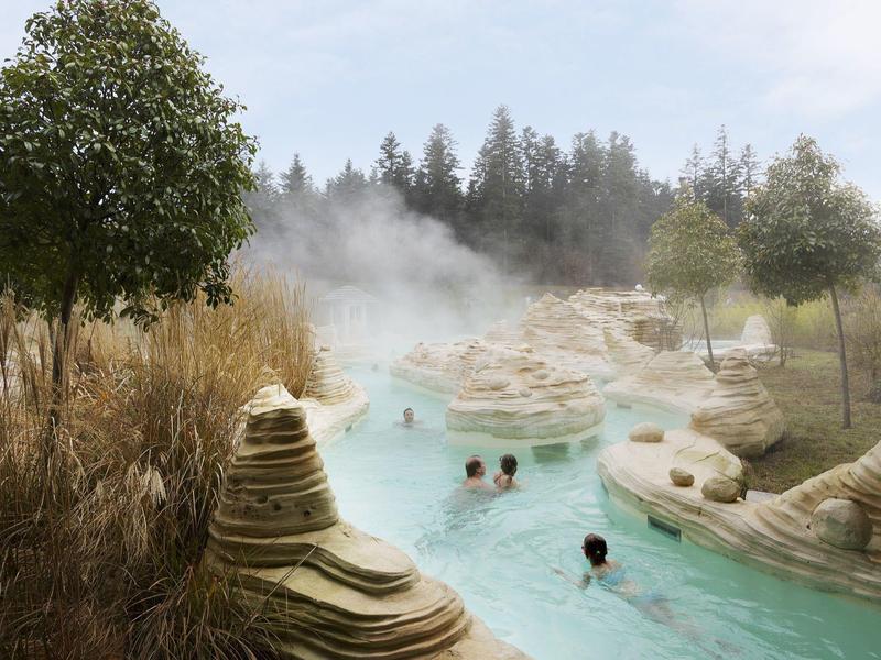 Warmwaterbron met stoom, omgeven door bomen en bezoekers in helder blauw water.