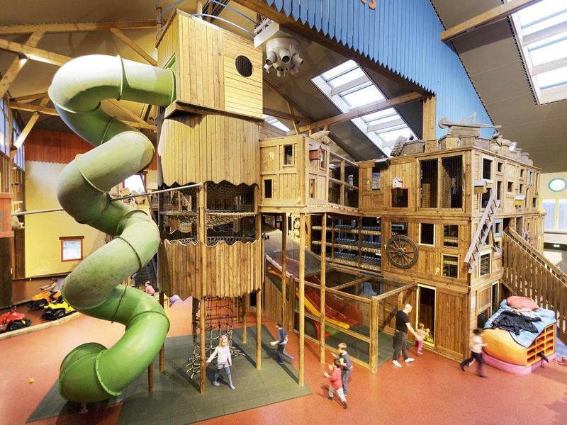 Groot indoor speelterrein met klimtoestellen en groene spiraalglijbaan in een hal.