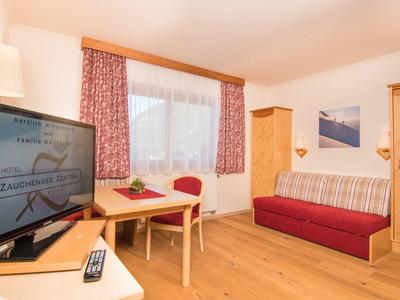 Gemütliches Wohnzimmer mit Sofa, Fernseher, Holztisch, Stühlen und Fenster mit roten Vorhängen.