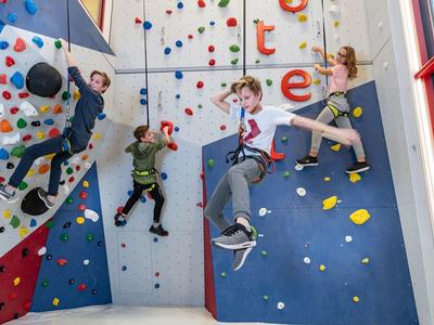 Vier Kinder klettern an einer bunten Kletterwand in einer hellen Halle mit großen Fenstern.