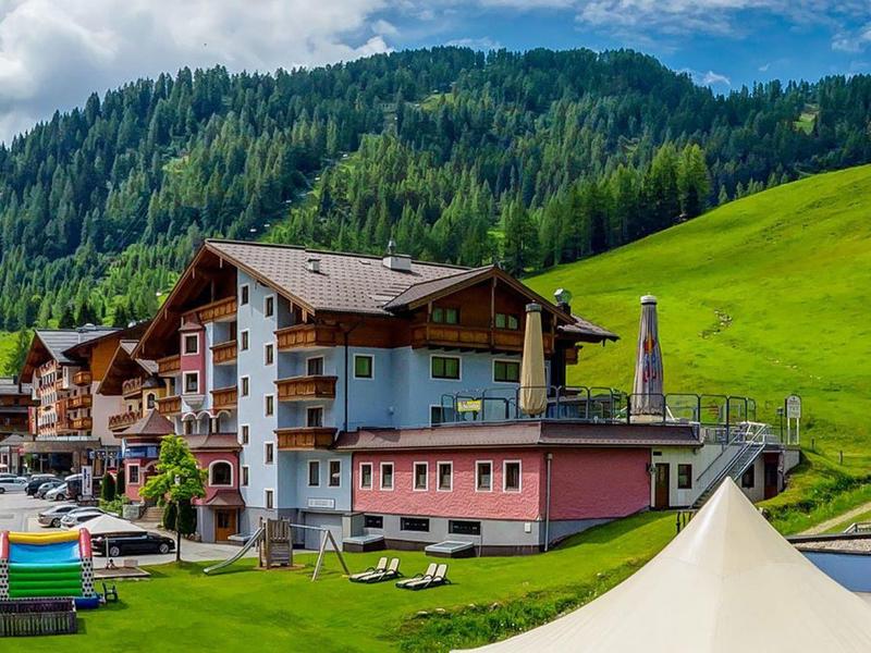 Alpines Dorf mit traditioneller Hütte, grünen Wiesen und bewaldeten Bergen im Hintergrund.
