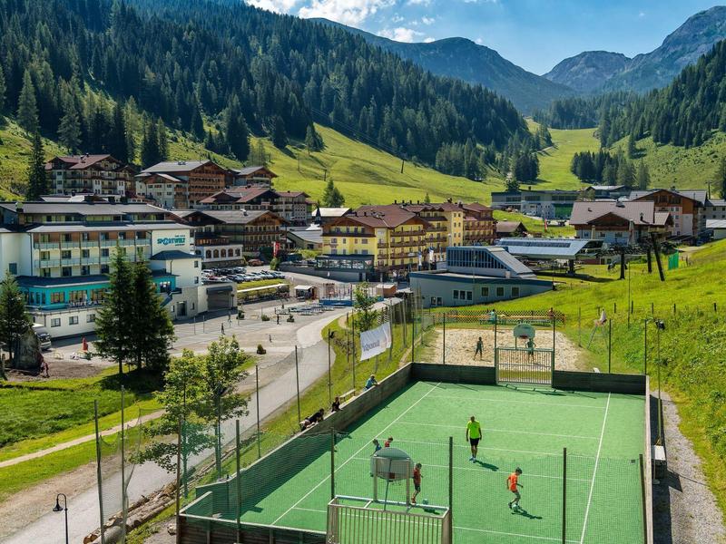 Dorf in Bergtal mit grünen Wiesen, Häusern und Tennisplatz im Vordergrund bei Sonnenschein.