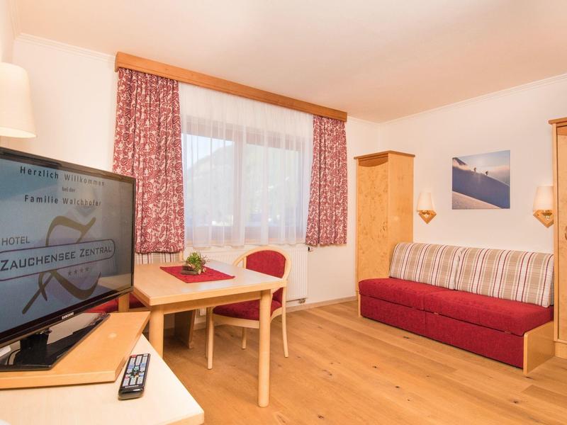 Gemütliches Wohnzimmer mit Sofa, Fernseher, Holztisch, Stühlen und Fenster mit roten Vorhängen.