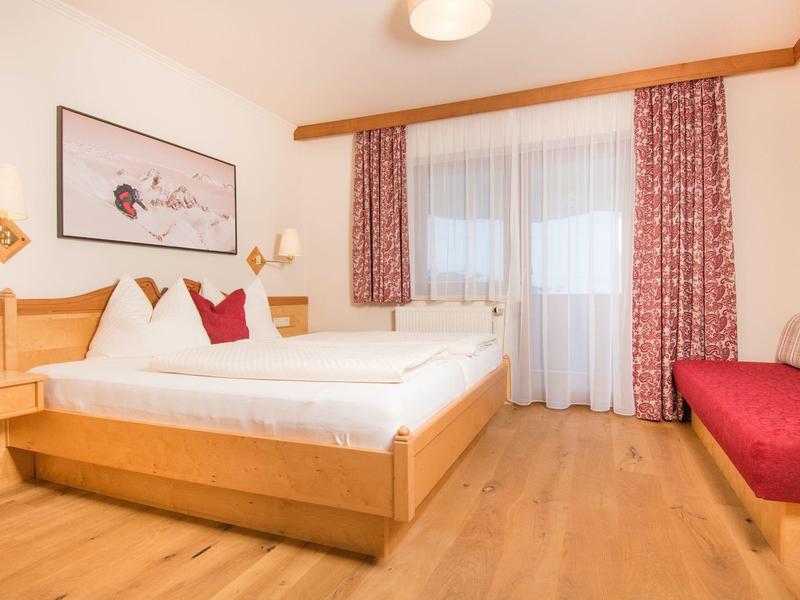 Helles Schlafzimmer mit Holzmöbeln, Doppelbett, roten Vorhängen und Sofa neben Fenstern.
