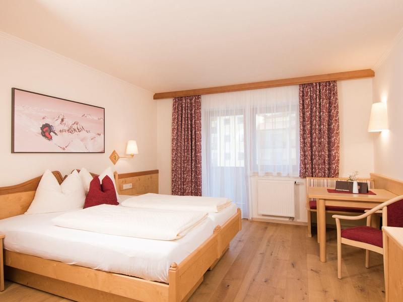 Helles Schlafzimmer mit Doppelbett, Holzmöbeln, Schreibtisch und großen Fenstern mit Vorhängen.