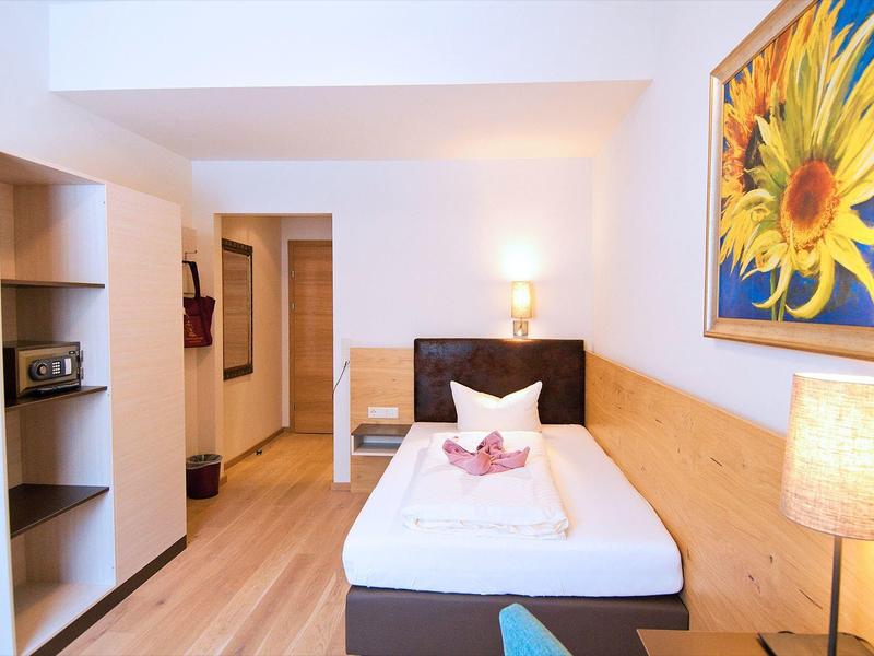 Kleines, helles Einzelzimmer mit Holzboden, gemütlichem Bett, Lampe und Sonnenblumenbild an der Wand.