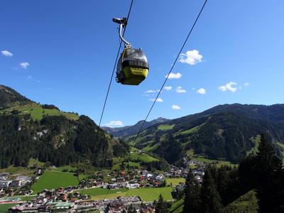 Gelbe Gondel-Seilbahn über grünem Tal mit Dorf und bewaldeten Bergen unter blauem Himmel.
