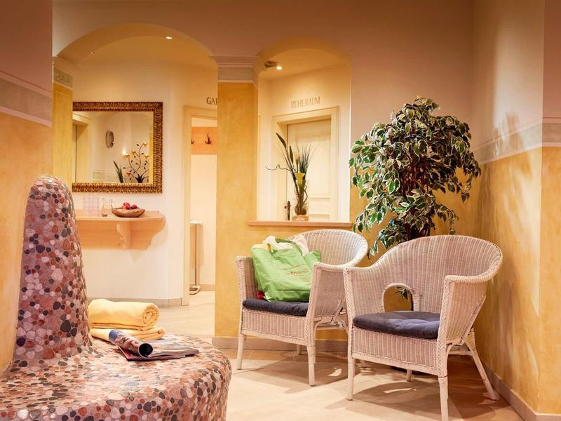 Gemütlicher Wartebereich mit Korbstühlen, grünem Kissen, Pflanzen und warmen Farben.