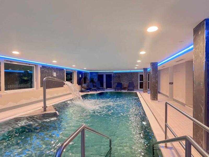 Moderner Innenpool mit blauen LED-Lichtern, Wasserfontäne und Metallgeländern in hellem Raum.