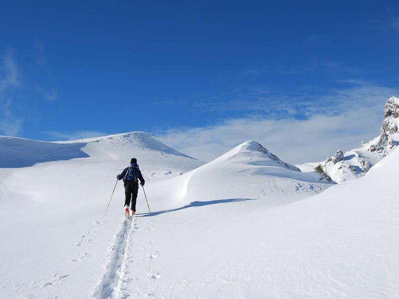 Eine einzelne Person wandert mit Skiern durch eine verschneite Berglandschaft unter blauem Himmel.