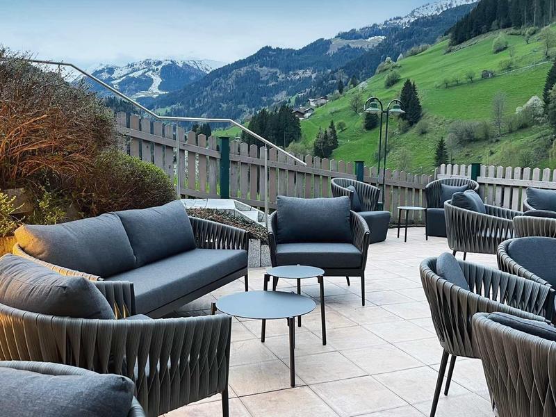 Terrasse mit modernen grauen Sitzmöbeln und Tischen, Berglandschaft mit grünen Wiesen im Hintergrund.
