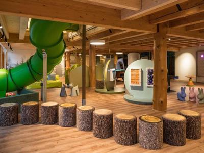 Kinderspielbereich in natürlichem Holzdielenraum mit grünem Röhrenrutsche und Baumstamm-Hockern.