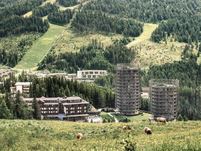 Mehrere Gebäude und Türme vor bewaldeten Hügeln mit grasbewachsenem Hang im Vordergrund.