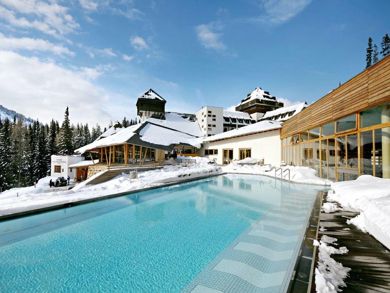 Moderner Außenpool vor einem Hotel in verschneiter Berglandschaft unter blauem Himmel.