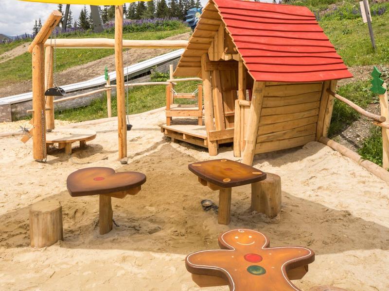 Kinderspielbereich mit kleinem Holzhaus, Tisch und Sitzgelegenheiten in Sandfläche und Sonnenschutz