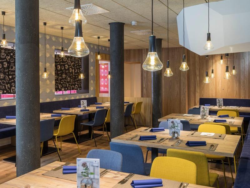 Modernes Restaurant mit Holztischen, gelben und blauen Stühlen und hängenden Glühbirnenlampen.