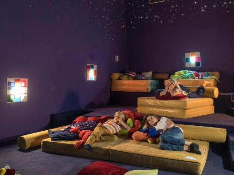 Kinder liegen auf bunten Matratzen in einem abgedunkelten Raum mit Sternenlichtprojektoren.