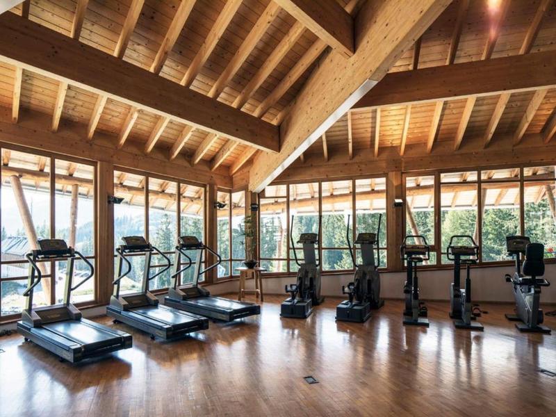 Großer Fitnessraum mit Holzboden, Holzbalkendecke und mehreren Trainingsgeräten vor großen Fenstern.