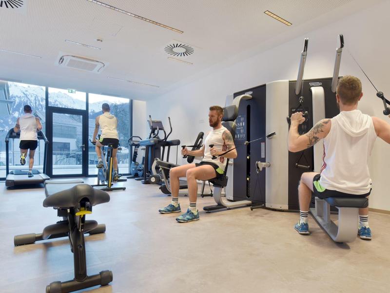 Helles Fitnessstudio mit drei Männern, die an verschiedenen Geräten trainieren, große Fenster.