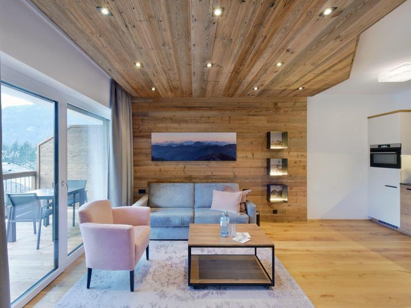 Modernes Wohnzimmer mit Holzboden, grauem Sofa, rosa Sessel, großem Fenster und offener Küche.