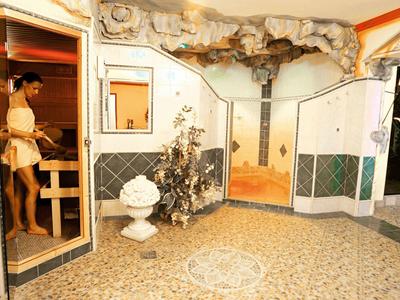 Innenhof mit Steinboden, dekorativer Wandmalerei, Springbrunnen und mehreren Türen, eine Frau betritt eine Sauna.