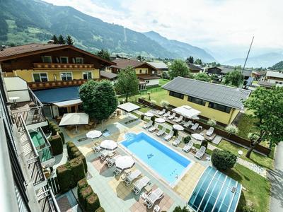 Blick auf einen Hotelgarten mit blauem Pool, Liegestühlen und umgeben von Bergen und Häusern.