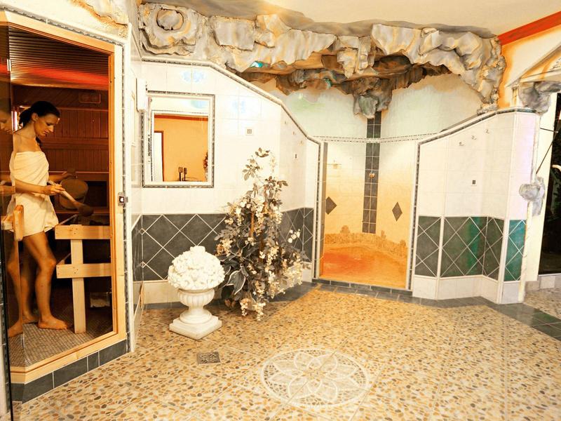 Innenhof mit Steinboden, dekorativer Wandmalerei, Springbrunnen und mehreren Türen, eine Frau betritt eine Sauna.