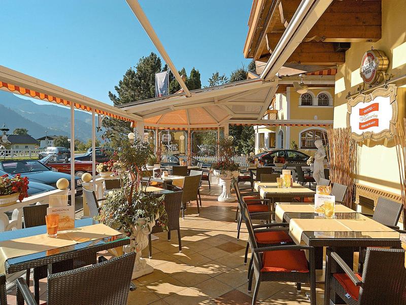 Sonnige Außenterrasse mit Holzdach, Tischen, Stühlen und Blumen vor einem Gasthaus.