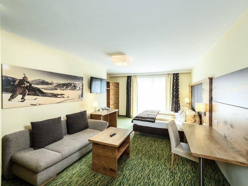 Helle Hotelzimmer mit grauem Sofa, Holztisch, Schreibtisch und Doppelbett am Fenster.