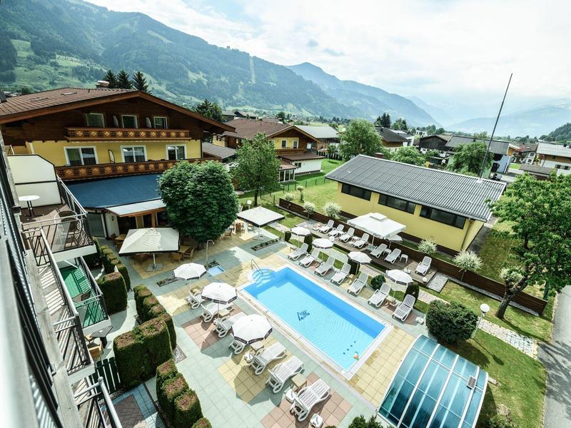 Blick auf einen Hotelgarten mit blauem Pool, Liegestühlen und umgeben von Bergen und Häusern.
