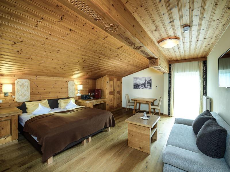 Gemütliches Schlafzimmer mit Holzwänden, Doppelbett, Sofa, Tisch und großem Fenster mit Vorhang.