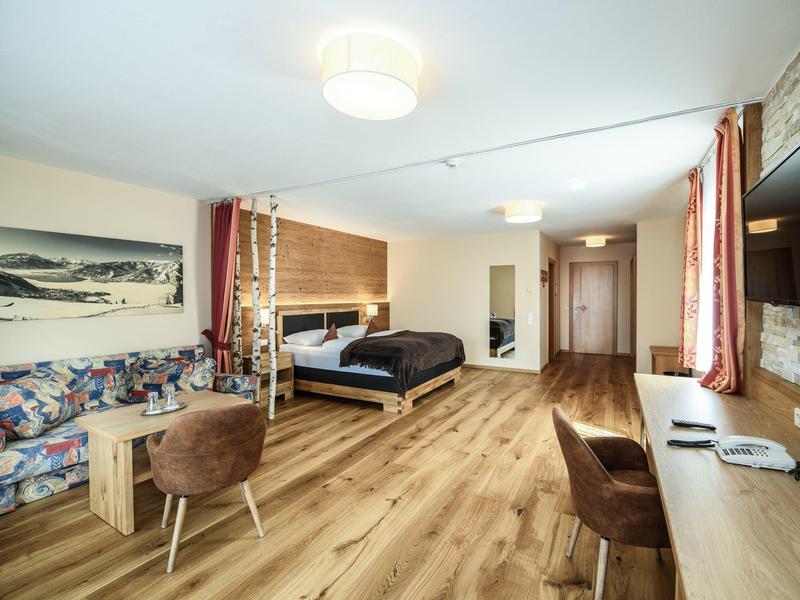 Helles, modernes Zimmer mit Holzboden, Doppelbett, Schreibtisch, zwei Sesseln und buntem Sofa.